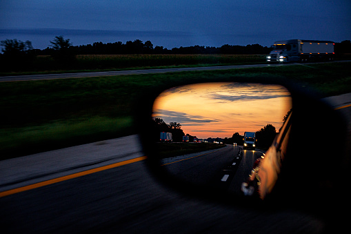Car driver point of view rear-view mirror reflection of sunset freight transportation heavy trucking traffic following and passing on a multiple lane highway expressway at dusk.