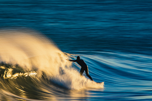Surfer catching a golden wave in strong wind reflecting a bright golden sunrise at the beach, Southern California, USA.