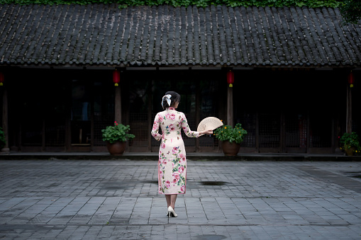 A beautiful woman in a cheongsam was standing in front of an old house with a fan in her hand