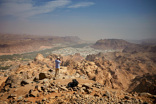 Full length rear view of mature man standing at rocky elevation and photographing Al-Ula Valley in northwestern Medina region.