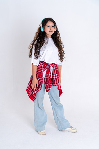 In full growth, young girl of caucasian appearance with long curly hair likes listen to music in headphones on white background. Brunette wearing white t-shirt, blue jeans and plaid red shirt .  Studio photo.