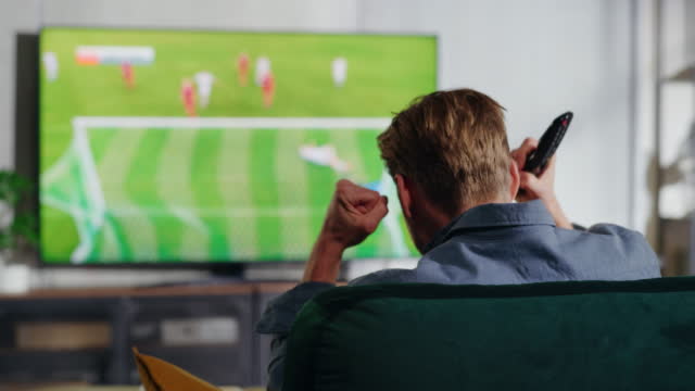 Male Soccer Fan Relaxes on a Couch, Watch a Sports Match at Home in Stylish Loft Apartment. Excited Young Man Cheer for Their Favorite Football Club, Celebrate Player Scoring a Goal.