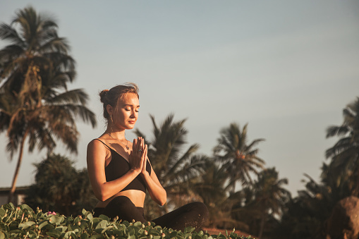 Young woman does yoga for healthy lifestyle on sea beach at palm trees background. Female performing sports exercises to restore strength and spirit. Yoga position on tropical climate