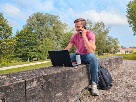 Mature caucasian male working with his laptop and smart phone. He is sitting on a bench in a pubic park.