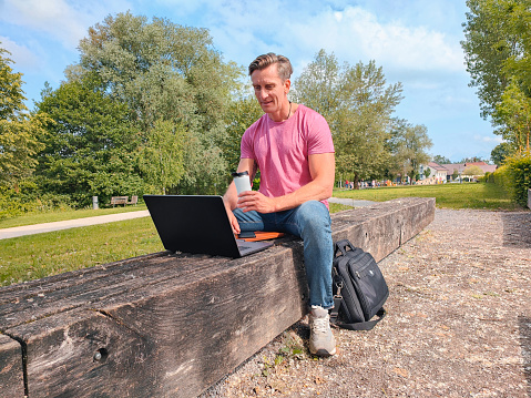 Mature caucasian male working with his laptop. He is sitting on a bench in a pubic park.