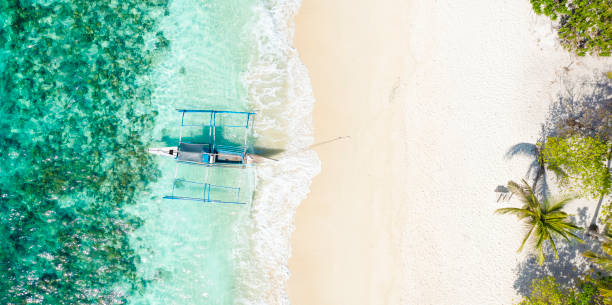View from above, stunning aerial view of a Bangka boat in front of a white sand beach bathed by a turquoise water. A Bangka is a double outrigger canoe native of the Philippines. Coron Island, Palawan, Philippines. View from above, stunning aerial view of a Bangka boat in front of a white sand beach bathed by a turquoise water. A Bangka is a double outrigger canoe native of the Philippines. Coron Island, Palawan, Philippines. outrigger stock pictures, royalty-free photos & images