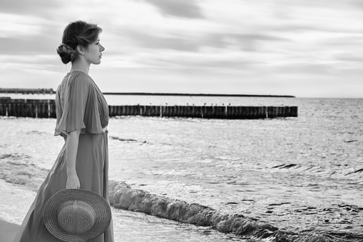 young luxury woman in dress with straw hat near sea looking away profile view, monochrome image