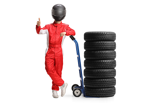 Full length portrait of a racer with a helmet leaning on a hand truck with tires and gesturing thumbs up isolated on white background