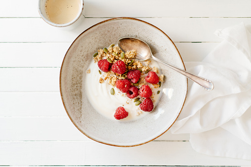 Granola with raspberries and yogurt. Beside the plate is a white napkin and a cup of coffee.