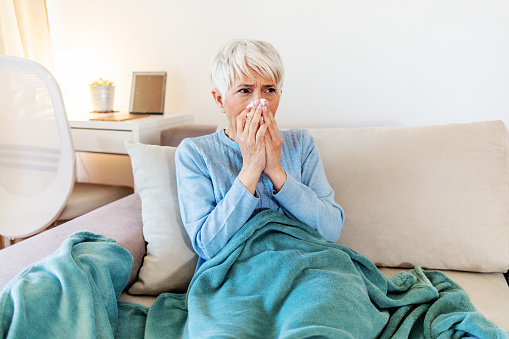 Mature woman with flu blowing nose at home. Close up elderly sick woman covered in blanket has cold blows her runny nose and suffers from flu condition.