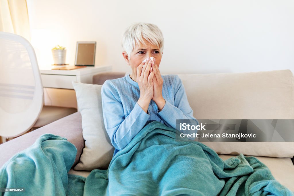 Shot of a senior woman blowing his nose with a tissue at home Mature woman with flu blowing nose at home. Close up elderly sick woman covered in blanket has cold blows her runny nose and suffers from flu condition. Cold And Flu Stock Photo