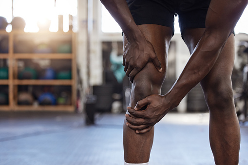 Closeup of one unknown african american athlete suffering from knee injury during workout in gym. Strong, fit, active black man feeling pain in leg joint during training and exercise at health club