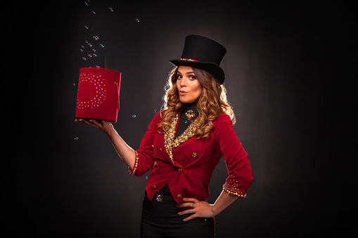 Female magician makes with soap bubbles show, an illusionist in theatrical clothes and hat, on black background. Woman actress in stage costume. Concept of theatrical performance and fun show