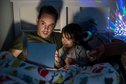 A close-up front view shot of a young boy wearing pyjamas lying in his bed with his father. His father is reading him a bedtime story out of a book.  There is a colourful night light projector that is projecting stars across the boys room.