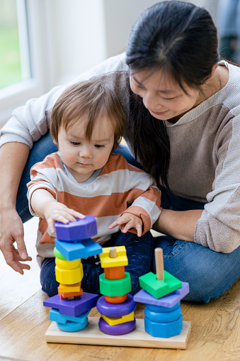 A close up front view shot of a mid-adult mother sitting on the floor of a living room with her young boy sitting with her, they are wearing casual clothing playing with toy building blocks together.