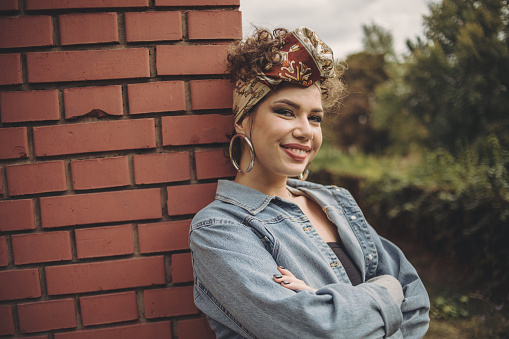 An urban young woman standing next to a brick wall, wearing denim has a turban on her head and a special urban style of dress.