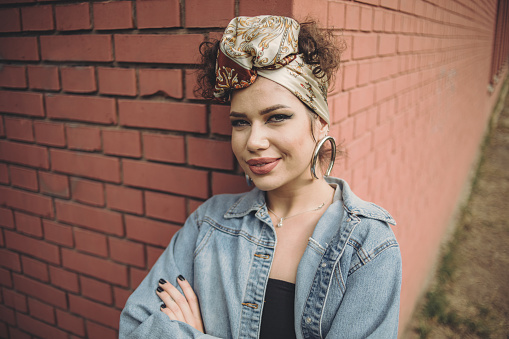 An urban young woman standing next to a brick wall, wearing denim has a turban on her head and a special urban style of dress.