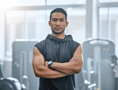 Portrait of one serious asian trainer alone in gym. Handsome focused coach standing with arms crossed after workout in health club. Young confident man in fitness centre for routine training exercise