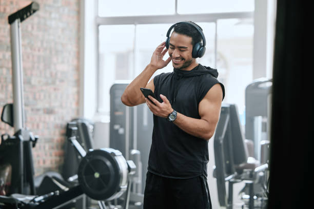 Smiling asian trainer alone in gym, listening to music on headphones from cellphone. Handsome coach standing in health club workout, selecting song from playlist on technology. Man in fitness centre Smiling asian trainer alone in gym, listening to music on headphones from cellphone. Handsome coach standing in health club workout, selecting song from playlist on technology. Man in fitness centre gym men africa muscular build stock pictures, royalty-free photos & images