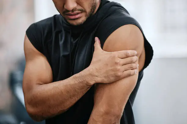 Photo of Unknown trainer alone in gym and suffering from shoulder injury. One coach standing and rubbing arm during workout in exercise health club. One man in fitness centre feeling pain in routine training