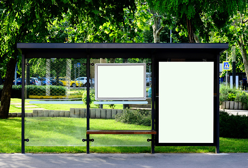 image composite of bus shelter at a bus stop. transparent clear display glass lightbox. aluminum frame structure. street perspective with trees. green background. empty white poster ad and advertising display glass.