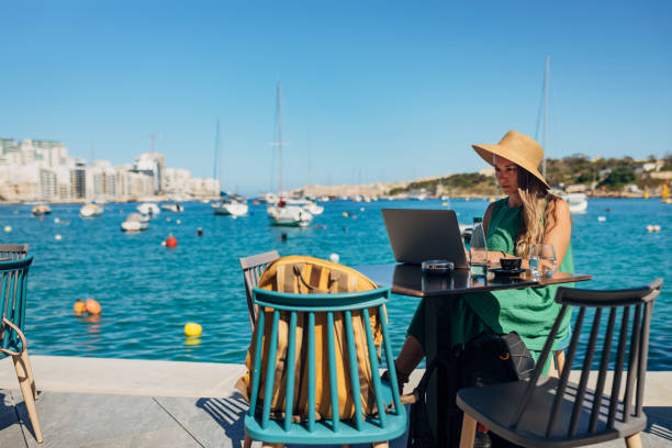 A business woman sitting in a coffee shop on the dock, not far from the sea and working on a laptop A woman with a straw hat on her head works remotely sitting in a coffee shop not far from the sea digital nomad stock pictures, royalty-free photos & images