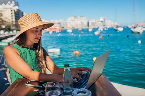 A female freelancer sits in a coffee shop not far from the harbor and uses a laptop to work remotely