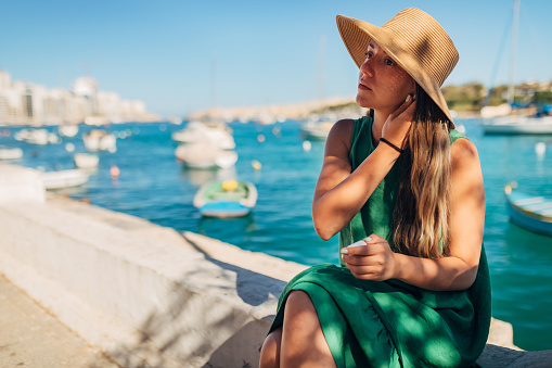 A millennial woman sitting on a dock not far from the sea puts on her wireless headphones