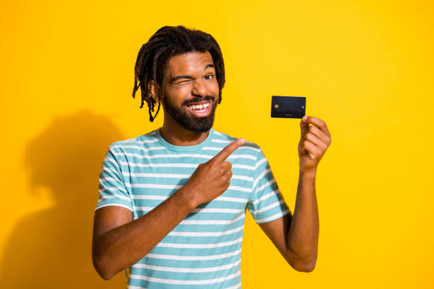 Photo portrait of winking black skin guy pointing finger at holding credit card in one hand isolated on vivid yellow colored background Photo portrait of winking black skin guy pointing finger at holding credit card in one hand isolated on vivid yellow colored background young man wink stock pictures, royalty-free photos & images