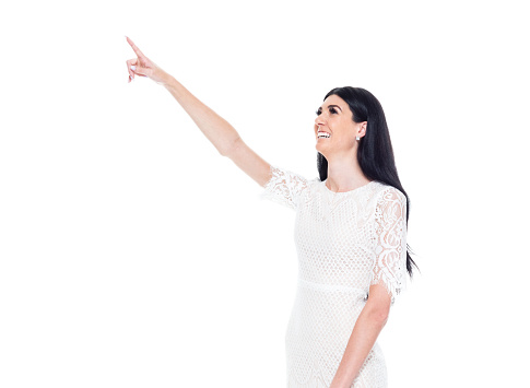 Side view of aged 20-29 years old who is beautiful caucasian young women standing in front of white background wearing dress who is cheerful who is pointing