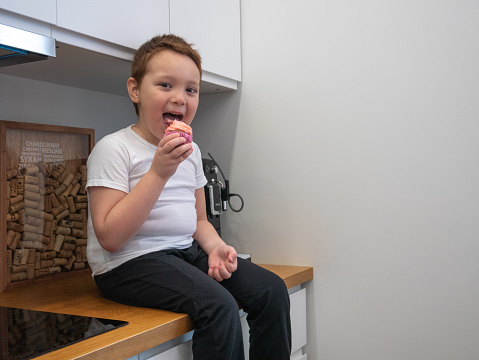 A little boy is sitting on the kitchen table, licking cream from a cupcake, smiling, eating deliciously, looking into the frame.