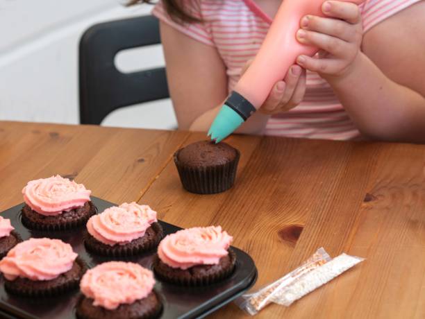 istock A child in the kitchen squeezes a cream from a confectionery bag on a cupcake. A baking sheet with ready -made cupcakes with pink cream on a wooden table 1401929544
