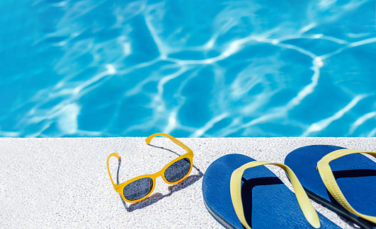 back view of flip flops and goggles on the white border of a swimming pool with bluish water in the background. vacation and summer concept.