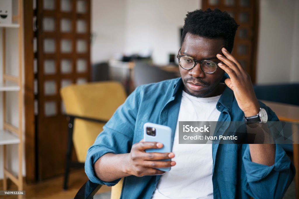Reading about bad news using a smart phone Young man sitting at home, feeling depressed and trying to contemplate bad news he is reading online using a smart phone Using Phone Stock Photo