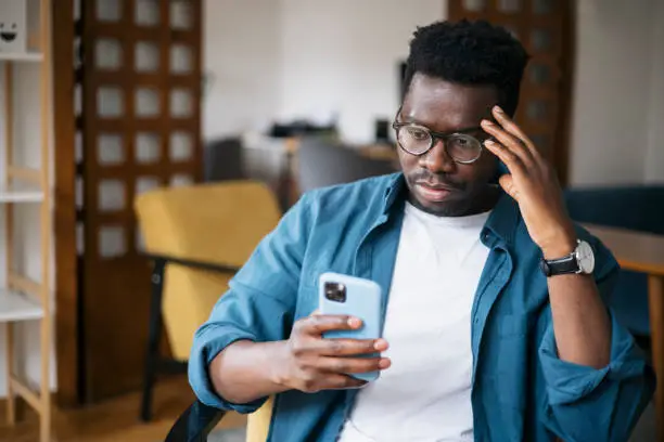 Young man sitting at home, feeling depressed and trying to contemplate bad news he is reading online using a smart phone