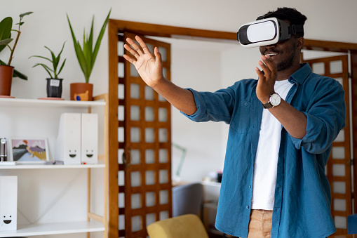 Male gamer standing in a living room at home, having VR googles on and playing games while holding his hands up in front of him
