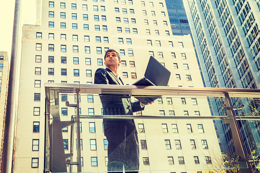 Way to Success. East Indian American student studying in New York, wearing black suit, standing by railing in business district, working on laptop computer, looking up, thinking.