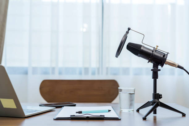 Studio microphone for recording podcasts sits on a table with a laptop, recording paper, pen and a glass of water.