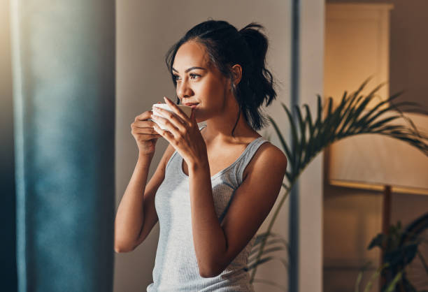 朝食に温かいコーヒーを楽しむ美しい若いヒスパニック系女性。アパートの窓からの眺めを眺めながらお茶を飲む混血女性 - drinking ストックフォトと画像