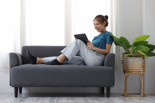 Cheerful Asian young woman lifestyle while sitting on sofa at home using tablet and having fun.