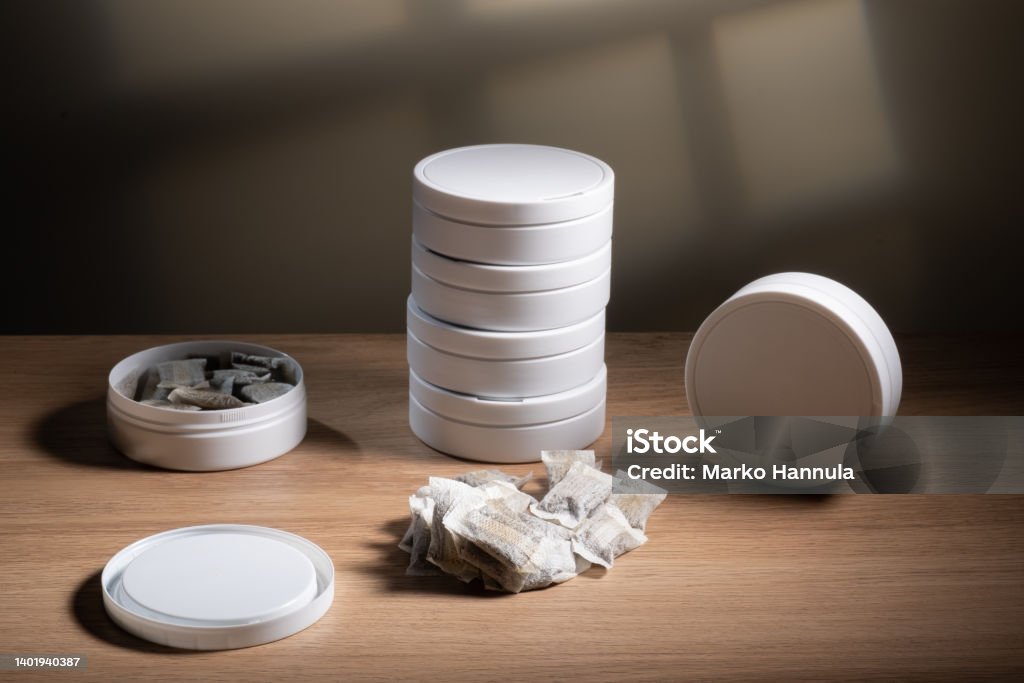 Studio shot of white swedish smokeless tobacco cans and portion snus pouches on a wooden table. Helsinki / Finland - JUNE 9, 2022: Studio shot of white swedish smokeless tobacco cans and portion snus pouches on a wooden table. Addiction Stock Photo