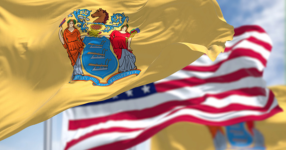 The New Jersey state flag waving along with the national flag of the United States of America. In the background there is a clear sky. New Jersey s a state in the Northeastern regions of the US