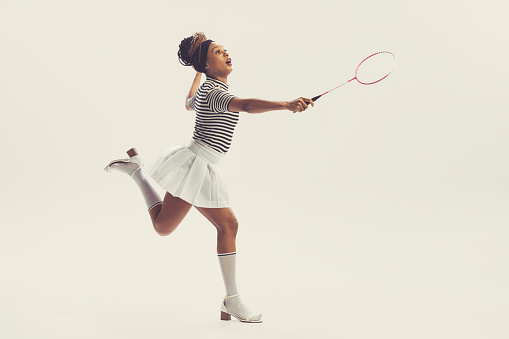 Leisure activities. Young happy beautiful girl in retro, 70s, 80s fashion style clothes, vintage outfit playing badminton isolated on white background. Concept of culture, art, music, fashion style.