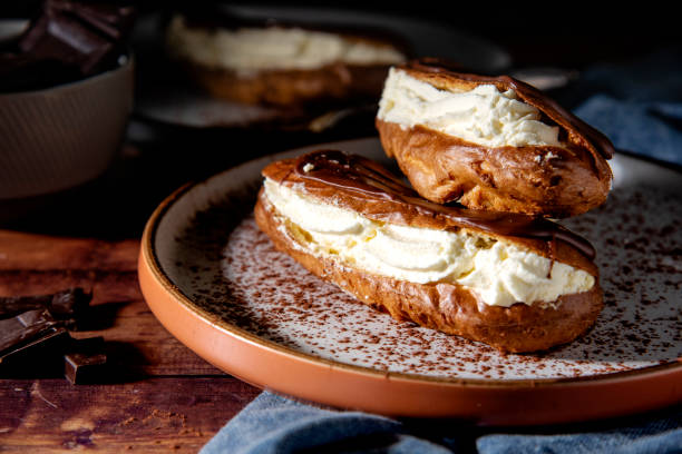 Homemade Chocolate eclairs on a ceramic plate with chocolate pieces on the side and another chocolate eclair in the background. The photograph is dark and moody.