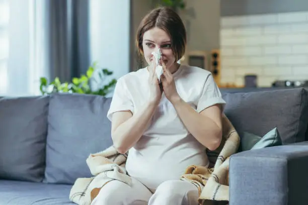 Photo of Disease. A pregnant woman is ill, has a runny nose, wipes her nose from the common cold with a napkin. Feels bad. Sitting at home in home clothes on the couch