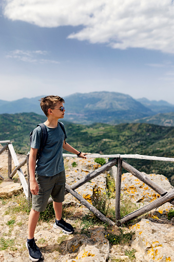 Teenage boy hiking in Sicilian mountains. He is standing on top of hill near town of Pollina.\nSunny spring day in Italy, Sicily.\nCanon R5