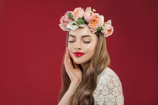 Beautiful young woman with healthy clean skin in summer rose wreath on red wall background