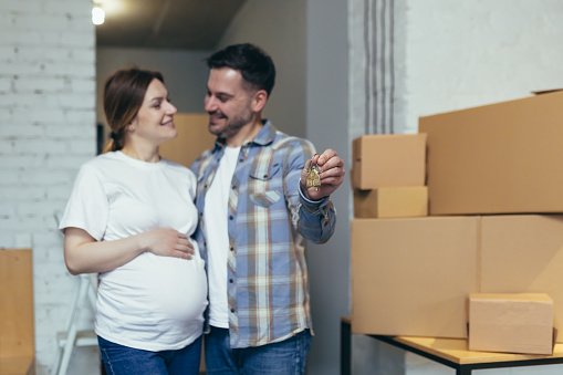 Young happy family expecting a baby. Pregnant woman and husband moved to a new house, apartment. Unpack boxes with things. Hugging, smiling
