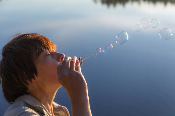 Caucasian young adult woman blowing soap bubbles outdoors near tall buildings, holding bubble wand and container, looking at bubbles floating in urban environment