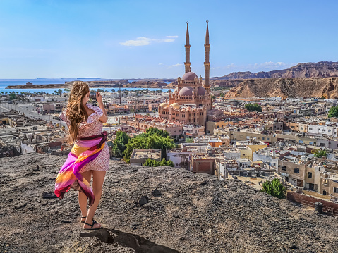 Female tourist shooting Al Sahaba Mosque in Sharm El Sheikh from the top of the cliff - view from the back. Young woman with a camera in her hands on the backdrop of a panorama of the Egyptian city